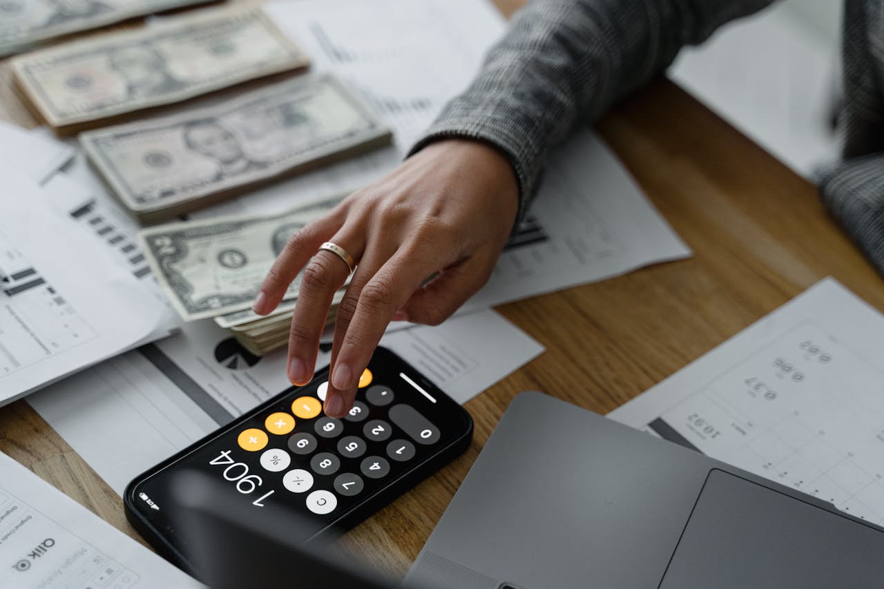 Close-up of a person using a smartphone calculator amid money and financial documents on a wooden table.